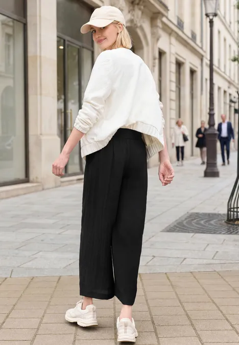 Eine Frau in weißer Jacke, Cap und schwarzen Hosen spaziert lächelnd durch die Einkaufspassage.