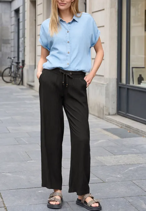 Eine Frau in heller Bluse, weiten schwarzen Hosen und Sandalen schlendert vor einem Stadthaus.