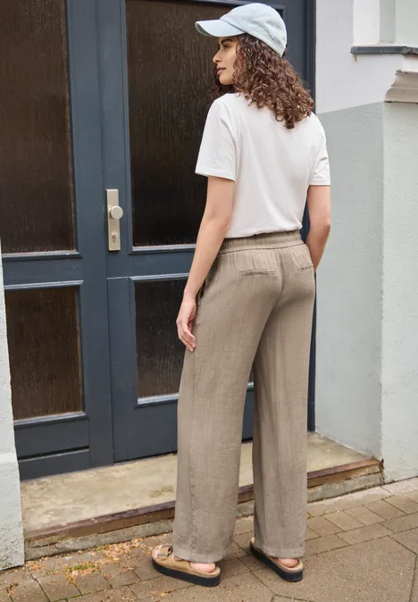 Straight Leg Hose mit Crinkle Struktur dark stone sand Frau in weißem T-Shirt, Cap, beigen Hosen und Sandalen lehnt am Hauseingang.