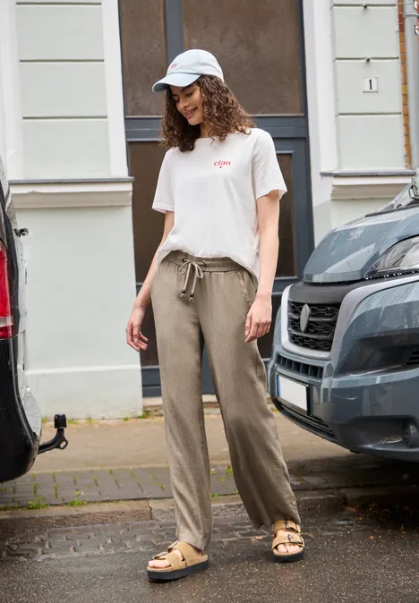 Straight Leg Hose mit Crinkle Struktur dark stone sand Junge Frau mit weißer Kappe, T-Shirt und weiten Stoffhosen steht lässig zwischen Autos auf dem Gehweg.