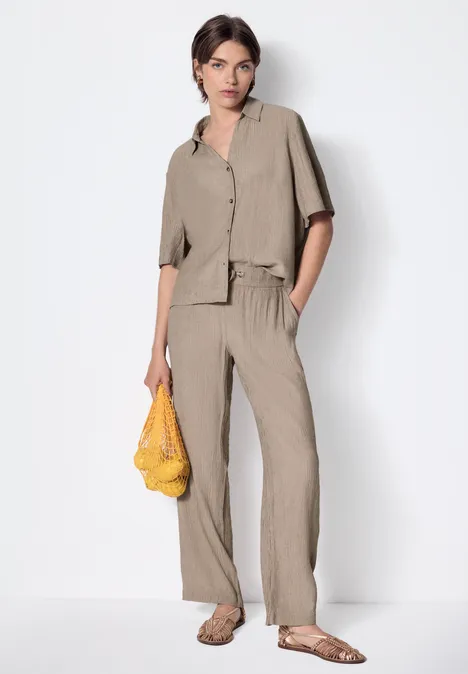 Straight Leg Hose mit Crinkle Struktur dark stone sand Frau in beiger Hemd-Hose-Kombi mit gelber Netztasche vor weißem Hintergrund.