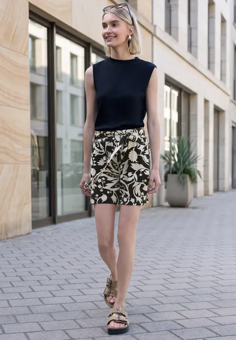 Eine Frau trägt schwarzen ärmellosen Top, gemusterte Shorts und Sandalen, geht entlang einer modernen Steinfassade.