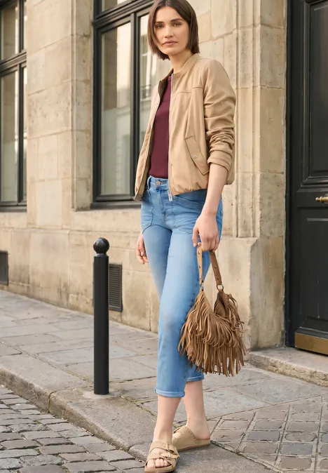 Vrouw in casual outfit met beige jasje, bordeaux top, blauwe jeans en franjes tas op kasseienstraat.