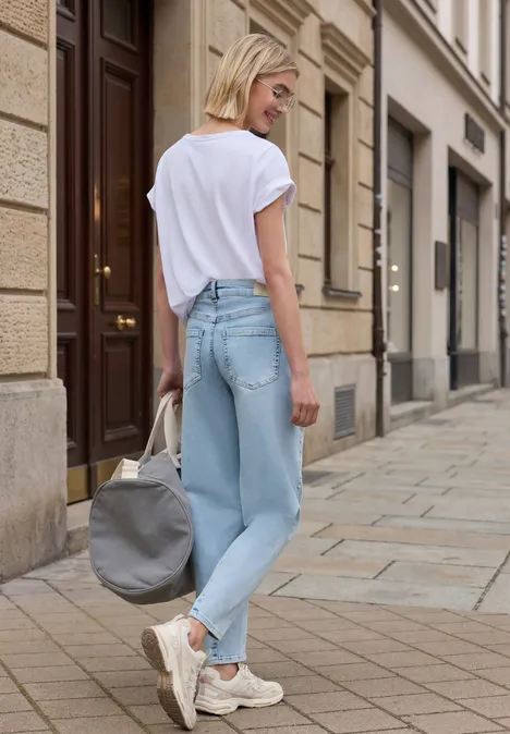 Eine junge Frau mit hellblauer Jeans, weißem T-Shirt und Sneakers geht mit grauer Tasche eine Straße entlang.