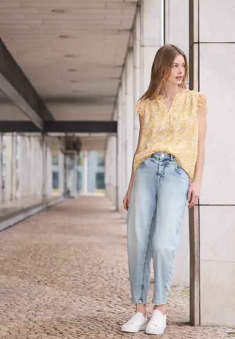 Frau in gelb geblümter Bluse, hellen Jeans und weißen Turnschuhen lehnt an einer Säule in modernem Durchgang.