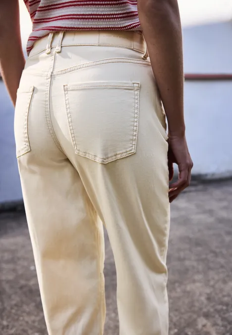 Vrouw van achteren in crème broek met zakken en gestreept topje, geleund tegen reling.
