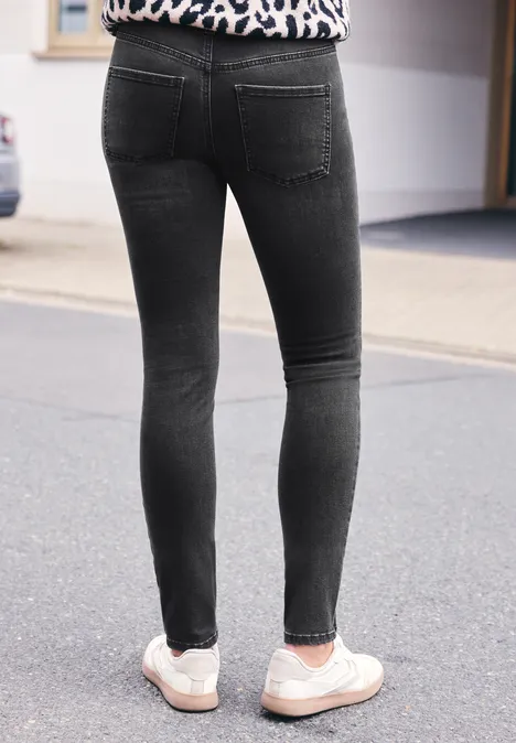 Mid Waist Skinny Leg Jeans im Skinny Fit black washed Eine Person steht auf dem Bürgersteig, trägt schwarze Skinny-Jeans und weiße Sneaker.