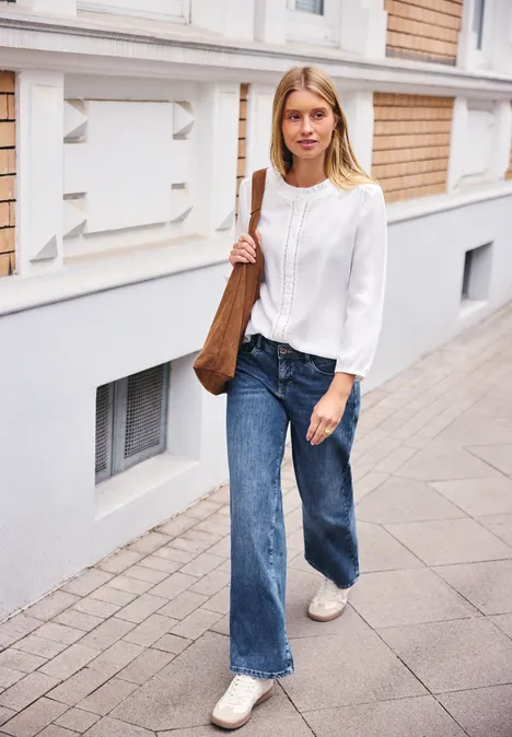 Mid Waist Wide Leg Jeans im Loose Fit bright mid blue washed Eine junge Frau in weißer Bluse, blauen Jeans und brauner Tasche geht auf dem Gehweg an einem Gebäude vorbei.