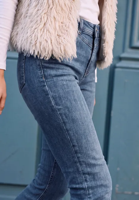 Eng anliegende blaue Jeans und weiße flauschige Weste vor einer türkisfarbenen Tür.