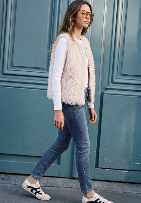 Frau in Jeans, weißem Langarmshirt, flauschiger Weste und Sneakers vor einer Haustür.