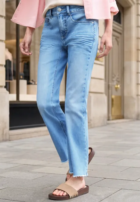 Frau trägt lockere blaue Jeans und Sandalen, urbane Straßenszenerie im Hintergrund.