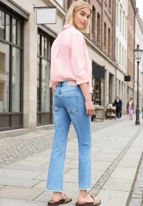 Eine Frau in rosa Bluse und Jeans steht auf einem Gehweg vor Geschäften.