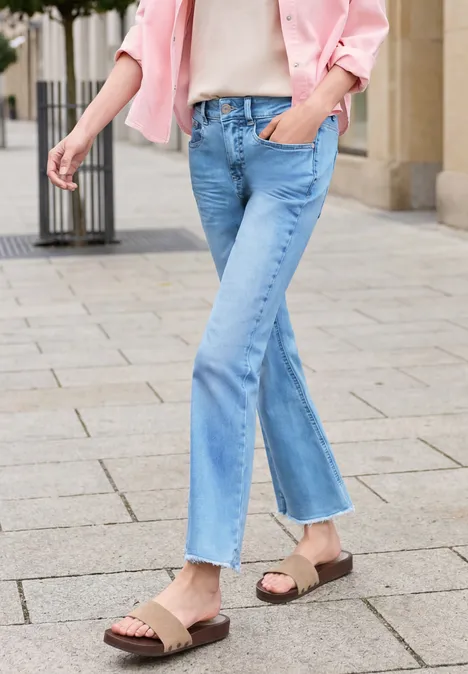 Person auf Gehweg in hellblauen, leicht ausgestellten Jeans, rosa Jacke und braunen Sandalen.