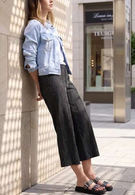 Frau in heller Jeansjacke und weiten schwarzen Hosen lehnt vor Schaufenster und gemusterten Säulen.