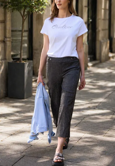 Frau spaziert in weißem T-Shirt, dunklen Hosen und mit hellblauem Blazer in der Hand über den Bürgersteig.