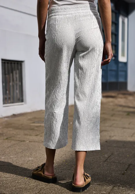 Rückenansicht einer Person in hellen, strukturierten, knöchellangen Hosen und flachen Sandalen auf dem Gehweg