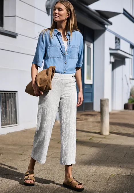 Frau in Jeanshemd, weißen Leinenhosen und Keilabsatz-Sandalen geht mit Strohhut spazieren.