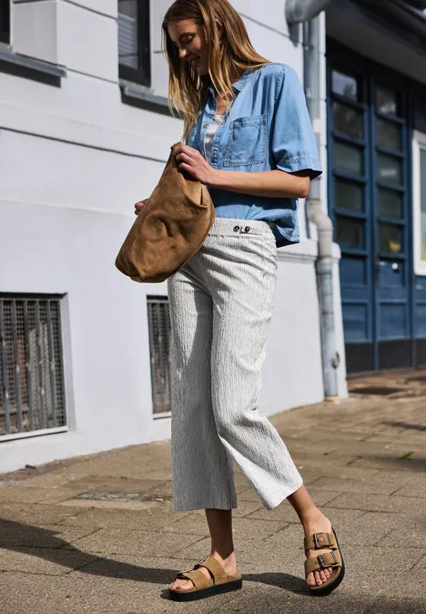 Frau in Jeanshemd und weißen Hosen mit brauner Tasche und Sandalen vor Hausfassade.