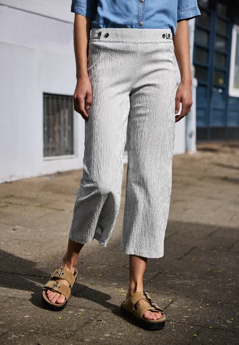 Beine einer Frau in gestreiften Leinen-Culottes und Sandalen, stehend auf einer Straße vor Hauswand.
