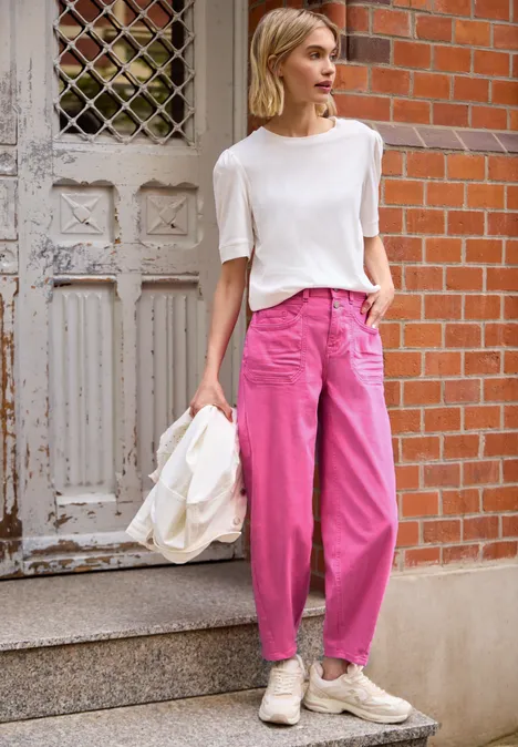 Barrel Leg Jeans im Loose Fit pink washed Frau in weißem T-Shirt, pinken, weiten Hosen und Sneakers, hält helle Jacke vor Tür und roter Backsteinwand.