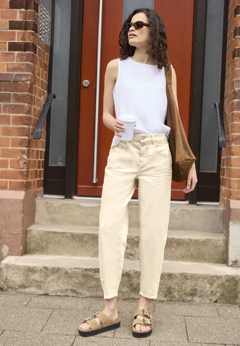 Barrel Leg Jeans im Loose Fit soft ecru Junge Frau vor roter Tür trägt weißen ärmellosen Oberteil, helle Hose und schwarze Sandalen mit Umhängetasche.