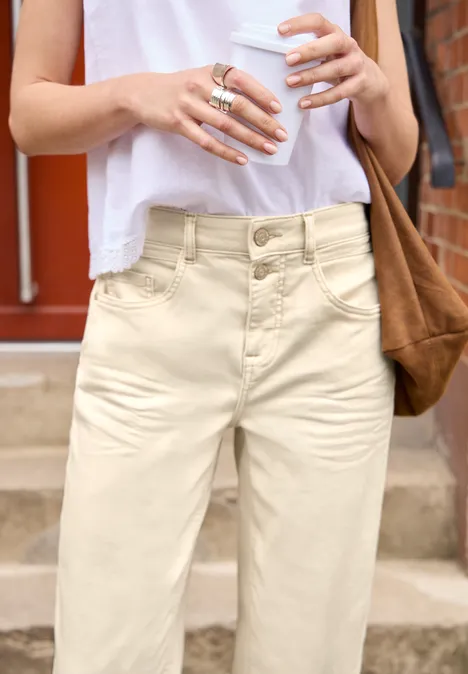 Barrel Leg Jeans im Loose Fit soft ecru Frau in weißem Top und cremefarbenen Hosen, mit brauner Schultertasche, hält einen Kaffeebecher und eine Wasserflasche.