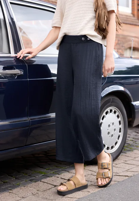 Vrouw in wijde donkerblauwe broek en sandalen staat naast een blauwe auto op kasseien.