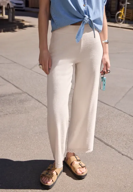 Wide Leg Hose mit Crochet-Detail vanilla sand Frau in weißen, weiten Hosen, blauem Oberteil und Birkenstock-Sandalen steht auf gepflastertem Gehweg.