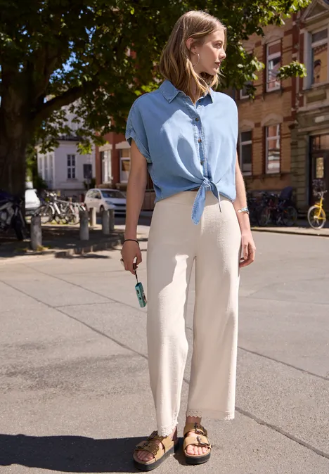 Wide Leg Hose mit Crochet-Detail vanilla sand Frau steht auf gepflastertem Platz, trägt hellblaue geknotete Bluse, helle weite Hose und Sandalen, hält Sonnenbrille.