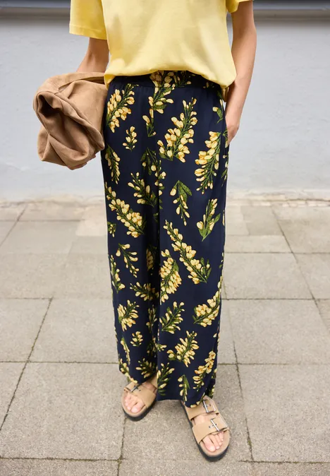 Femme portant un pantalon ample noir à motif floral jaune, avec un haut jaune et des sandales.