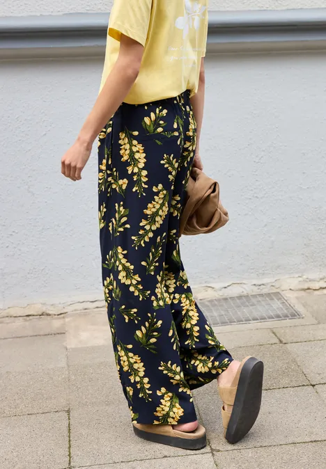 Personne marche sur le trottoir, t-shirt jaune, pantalon noir à fleurs jaunes et sandales plateformes.