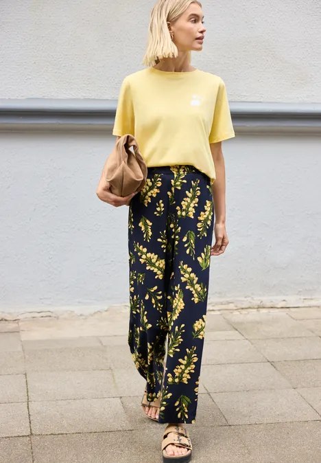 Une personne en t-shirt jaune et pantalon noir à motif floral jaune porte un sac marron en marchant.