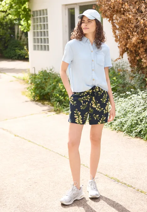 Une personne se tient sur un trottoir ensoleillé, porte une chemise bleu clair, un short à motif de feuilles et des baskets blanches.