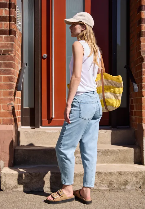 Barrel Leg Jeans im Loose Fit light blue random wash Eine Frau mit weißem Top, Cap, Jeans und Sandalen trägt eine gelb-weiße Tasche und steht vor einer Haustür.