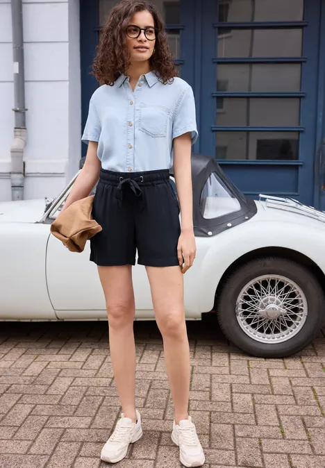 Vrouw in lichtblauw overhemd, zwarte short en witte sneakers voor een witte oldtimer.