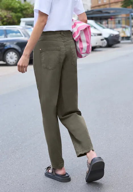 Person in olivgrünen Hosen und schwarzen Sandalen geht die Straße entlang und trägt eine rosa-weiß gestreifte Tasche.