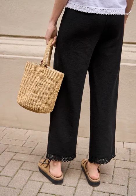 Wide Leg Hose mit Crochet-Detail Black Frau trägt weit geschnittene schwarze Caprihose mit Spitzenbesatz, Basttasche und Sandalen auf Bürgersteig.