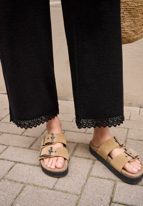 Wide Leg Hose mit Crochet-Detail Black Ein Paar Füße in goldenen Sandalen mit zwei Schnallen steht auf gepflastertem Boden.