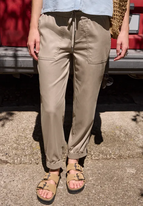 Slim Leg Joggpants in Cupro-Touch-Qualität dark stone sand Stehende Person trägt beigen Stoffhose mit Gummizug und beige Sandalen vor rotem Auto.