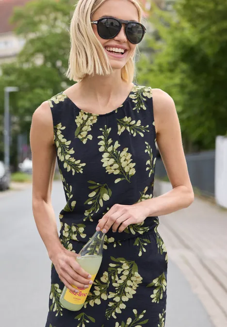 Frau mit schwarzem Top mit grünen Blättermuster hält eine Flasche und steht auf einem Gehweg.