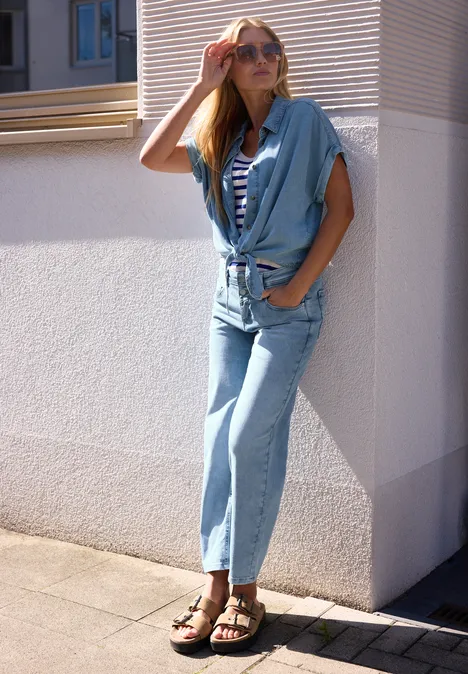 Femme debout contre un mur blanc, vêtue d'une combinaison en jean bleu clair, top rayé et sandales.