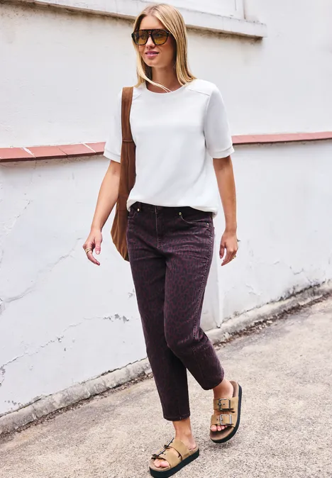 High Waist Barrel Leg Jeans mit Leo-Print burgundi printed wash Frau mit Sonnenbrille, weißer Bluse, gemusterten Hosen und Sandalen geht an einer weißen Hauswand entlang.