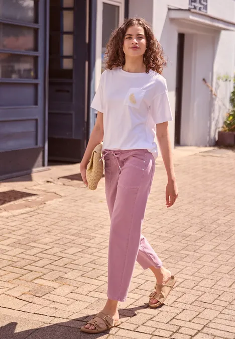 Eine junge Frau in weißem T-Shirt und lila Hose schlendert sommerlich durch die sonnige Gasse.
