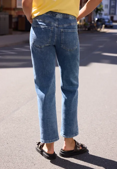 Rückansicht einer Person mit blauen Jeans und schwarzen Sandalen auf einer sonnigen Straße.
