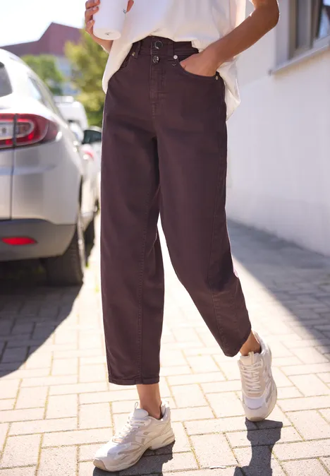 High Waist Barrel Leg Jeans w stylu Loose Fit burgundi color washed Osoba ma luźne, ciemne spodnie i białe sneakersy, stoi na chodniku obok samochodu.
