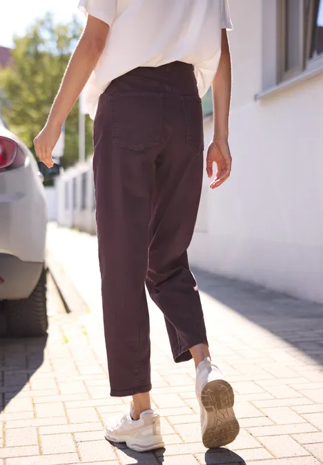 High Waist Barrel Leg Jeans w stylu Loose Fit burgundi color washed Osoba w białych trampkach i ciemnobrązowych spodniach idzie chodnikiem obok białego domu.