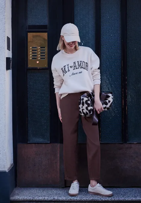High Waist Barrel Leg Jeans w stylu Loose Fit burgundi color washed Osoba w białej bluzie, beżowej czapce, brązowych spodniach i białych trampkach, trzyma torbę w motyw krowi na tle drzwi.