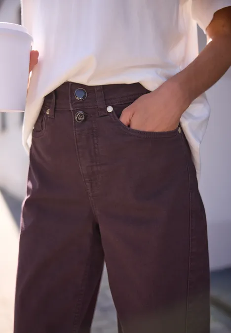 High Waist Barrel Leg Jeans w stylu Loose Fit burgundi color washed Zbliżenie osoby w brązowych dżinsach z ręką w kieszeni i kubkiem kawy.