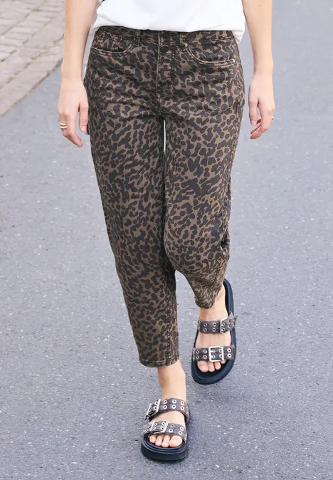Une personne marche sur un sol gris, portant un pantalon léopard brun et des sandales noires à boucles argentées.
