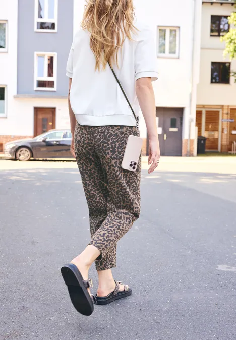 Femme marche dans la rue, porte un pantalon léopard, chemise blanche, sandales noires et tient un téléphone.